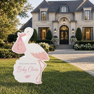 May include: A white and pink stork-shaped yard sign with the text "Welcome Home Baby Claire." The stork carries a pink bundle with the letter "C." The sign is placed on a green lawn in front of a large house.