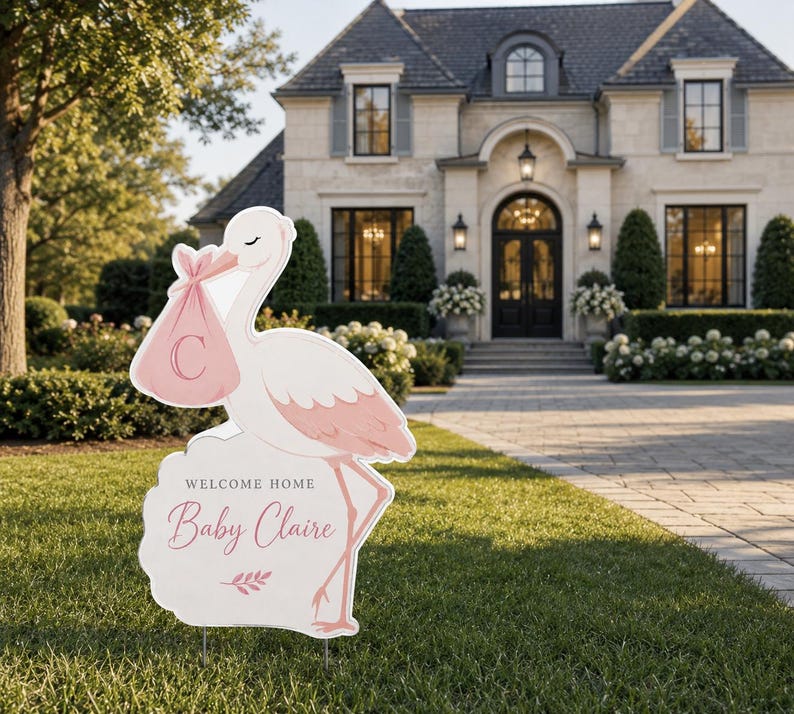 May include: A white and pink stork-shaped yard sign with the text "Welcome Home Baby Claire." The stork carries a pink bundle with the letter "C." The sign is placed on a green lawn in front of a large house.