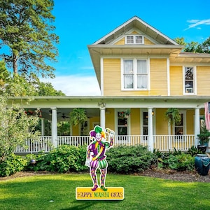May include: A yellow house with a white porch and a green lawn. There is a jester cutout in the front yard with the words "Happy Mardi Gras" written on the ground.