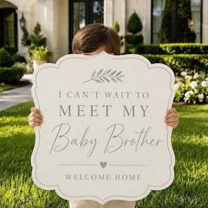 May include: A decorative sign with a scalloped edge, held by a person, features the text "I CAN'T WAIT TO MEET MY Baby Brother WELCOME HOME" in gray lettering. The sign has a light gray border and a sprig of leaves at the top. The background includes a house and green grass.
