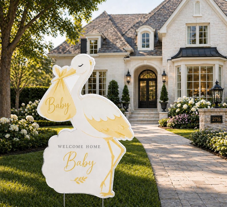 May include: A white and yellow stork-shaped yard sign with the word "Baby" on a pouch and on the sign. The sign reads "Welcome Home Baby" and is placed in a front yard.