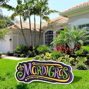 May include: A purple, green, and gold Mardi Gras sign with glitter and beads. The sign is in front of a house with a lush green lawn.