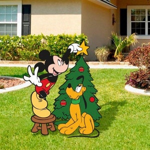 May include: Mickey Mouse and Pluto are decorating a Christmas tree with red and gold ornaments. Mickey is placing a star on top of the tree while Pluto watches.
