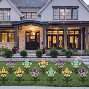 May include: A set of 12 purple, gold, and green fleur-de-lis yard decorations. The decorations are arranged in a row on a green lawn in front of a house.