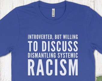 May include: Royal blue t-shirt with white text: "INTROVERTED, BUT WILLING TO DISCUSS DISMANTLING SYSTEMIC RACISM." A cup of coffee, a leather journal, and a sunflower are also in the image. The shirt is made of a soft material.