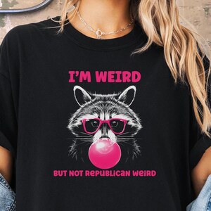 Peut inclure: T-shirt noir avec un graphique d'un raton laveur portant des lunettes de soleil et soufflant une bulle rose. Le texte "I'm Weird But Not Republican Weird" est imprim&eacute; sur le t-shirt.