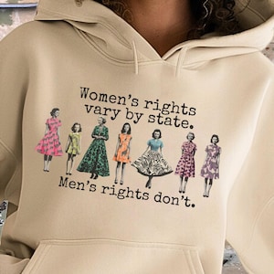 Puede incluir: Sudadera con capucha color arena con bolsillo delantero y capucha con cordón. El gráfico en la parte delantera presenta el texto "Women's rights vary by state. Men's rights don't." e ilustraciones de mujeres con vestidos.