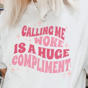 May include: White t-shirt with pink text that reads "Calling me woke is a huge compliment".