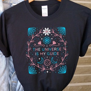 May include: Black t-shirt with a graphic design featuring a flower, moon, and stars in pink and blue. The text "The Universe is My Guide" is printed on the shirt.