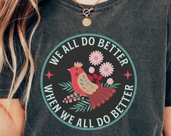 May include: A dark gray t-shirt with a circular graphic featuring a red bird with a crown, surrounded by flowers and leaves. The text "We all do better when we all do better" is written around the graphic.