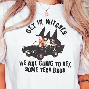 May include: White t-shirt with a black and white illustration of three witches driving a car. The text "Get in Witches We are going to hex some tech bros" is printed on the shirt.