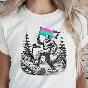 May include: Cream-colored t-shirt featuring a black and white illustration of a Bigfoot holding a flag with pink, green, and blue stripes. The design includes trees, a log, and mountains. The word "NATURAL" is printed at the bottom.