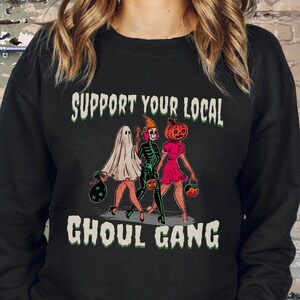 K&ouml;nnte beinhalten: Ein schwarzes Langarm-Sweatshirt mit einem Grafikdruck von drei Figuren in Halloween-Kost&uuml;men, darunter ein Geist, ein Skelett und eine Figur mit K&uuml;rbiskopf, sowie dem Text "SUPPORT YOUR LOCAL GHOUL GANG" in wei&szlig;er, verwaschener Schrift.