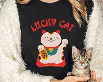 May include: A black t-shirt with the text "LUCKY CAT" in red and a cartoon image of a white Maneki-Neko, a Japanese lucky cat, with a gold bell and a red and gold base. The cat is holding up its right paw. A person is holding a tabby cat in their arms.