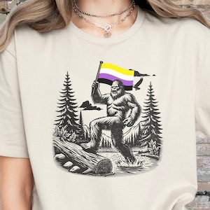 May include: A white t-shirt with a graphic of a Bigfoot holding a purple, yellow, and white flag. The flag is a symbol of the non-binary gender identity. The graphic is set against a backdrop of trees and mountains.