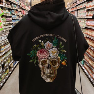 Puede incluir: Sudadera con capucha negra con un dise&ntilde;o de calavera y flores. El texto "Mantente amable en situaciones crueles" est&aacute; impreso en la parte posterior de la sudadera.