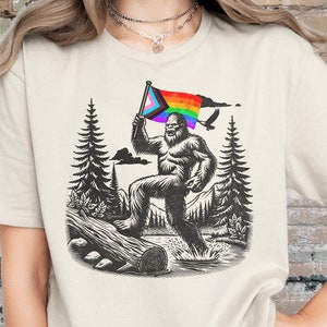 May include: A white t-shirt with a graphic of a Bigfoot holding a rainbow pride flag. The Bigfoot is walking across a log in a forest setting.