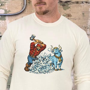 May include: Ivory long-sleeve shirt featuring a graphic of a lumberjack chopping ice with an axe, and a blue bull with a bell. The shirt is a solid ivory color.