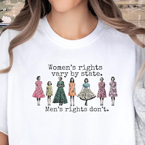 Puede incluir: Camiseta blanca con el texto "Women's rights vary by state. Men's rights don't." El dise&ntilde;o presenta ilustraciones de mujeres con vestidos. Una prenda casual y c&oacute;moda.