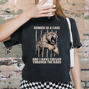 May include: Black t-shirt with a graphic of a leopard behind bars, with the text "GENDER IS A CAGE" above and "AND I HAVE CHEWED THROUGH THE BARS" below. The person is wearing a black beanie and checkered shorts.