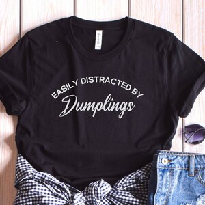 May include: Black t-shirt with white text that reads "Easily Distracted by Dumplings". The shirt is folded and displayed on a light wood background with a pair of sunglasses and a denim skirt.
