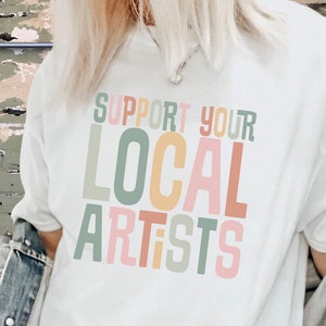 May include: A white t-shirt with the text "SUPPORT YOUR LOCAL ARTISTS" in colorful letters. The letters are stacked on top of each other and are in different shades of pink, green, yellow, and orange.