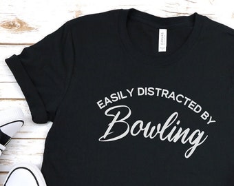 May include: Black t-shirt with white text that reads "Easily Distracted by Bowling". The shirt is laid flat on a white wooden surface with a pair of black sneakers and blue jeans.
