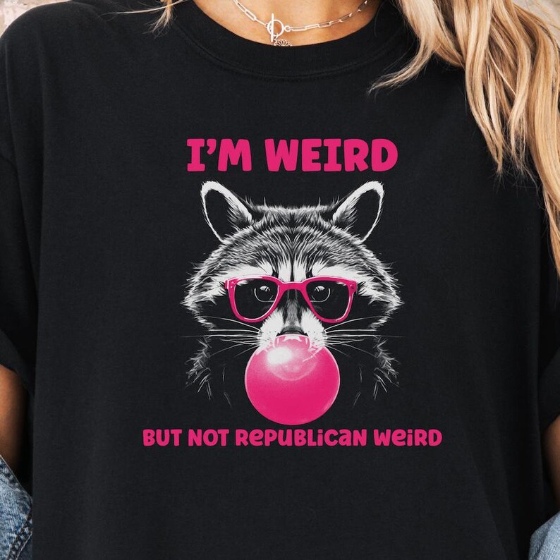 Peut inclure: T-shirt noir avec un graphique d'un raton laveur portant des lunettes de soleil roses et soufflant une bulle rose. Le texte "I'm Weird But Not Republican Weird" est imprim&eacute; sur le t-shirt.