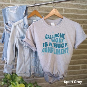 May include: A heather grey t-shirt with the text "Calling me woke is a huge compliment" in a blue and teal retro font. The shirt is on a hanger and has a distressed denim jacket and a pair of jeans in the background. The text "Sport Grey" is in the bottom right corner of the image.
