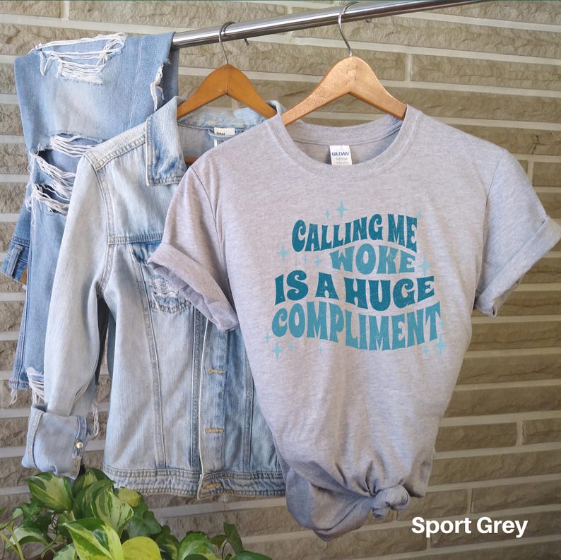 May include: A heather grey t-shirt with the text "Calling me woke is a huge compliment" in a blue and teal retro font. The shirt is on a hanger and has a distressed denim jacket and a pair of jeans in the background. The text "Sport Grey" is in the bottom right corner of the image.