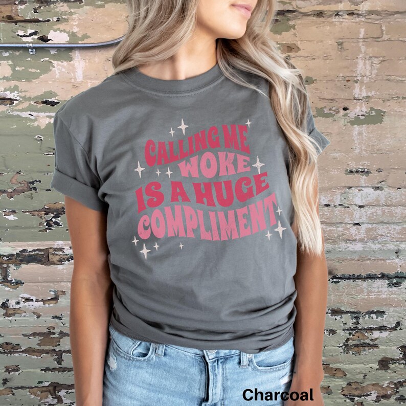 May include: A charcoal grey t-shirt with the text "Calling me woke is a huge compliment" in pink lettering with sparkling stars.
