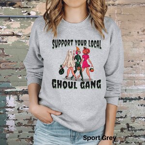 K&ouml;nnte beinhalten: Ein sportgraues Sweatshirt mit Rundhalsausschnitt und einem Grafikdruck von drei Figuren in Halloween-Kost&uuml;men sowie dem Text "SUPPORT YOUR LOCAL GHOUL GANG" in schwarzer und gr&uuml;ner Schrift. Das Motiv zeigt ein Gespenst, ein Skelett und eine K&uuml;rbisfigur.