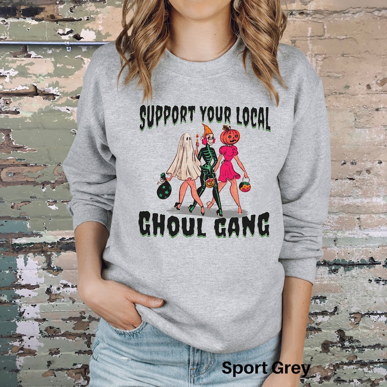 K&ouml;nnte beinhalten: Ein sportgraues Sweatshirt mit Rundhalsausschnitt und einem Grafikdruck von drei Figuren in Halloween-Kost&uuml;men sowie dem Text "SUPPORT YOUR LOCAL GHOUL GANG" in schwarzer und gr&uuml;ner Schrift. Das Motiv zeigt ein Gespenst, ein Skelett und eine K&uuml;rbisfigur.