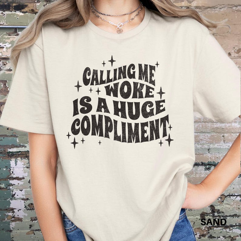 May include: A white t-shirt with a black graphic that says "Calling me woke is a huge compliment" with stars around the text.