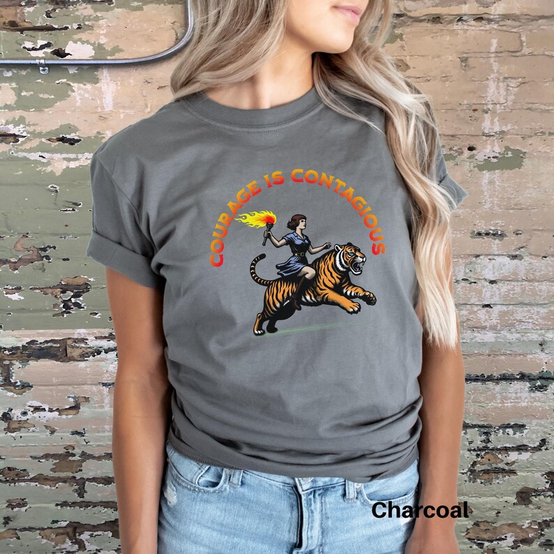 K&ouml;nnte beinhalten: Ein anthrazitfarbenes T-Shirt mit einem Motiv einer Frau, die auf einem Tiger mit einer brennenden Fackel reitet. Der Text "Courage is Contagious" ist in einem Kreis um das Motiv geschrieben.