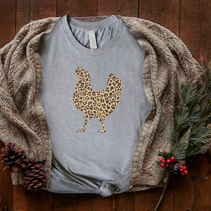 May include: A gray t-shirt with a leopard print chicken design. The shirt is laid out on a wooden surface with a brown knitted cardigan and a sprig of greenery.