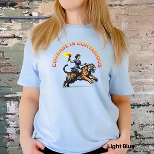 K&ouml;nnte beinhalten: Hellblaues T-Shirt mit einem Motiv einer Frau, die auf einem Tiger reitet. Der Text "COURAGE IS CONTAGIOUS" ist in einem Kreis um das Motiv gedruckt.