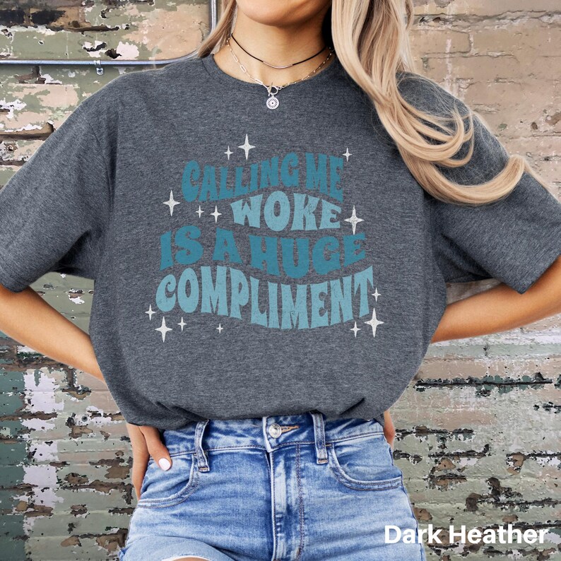May include: A dark heather grey t-shirt with the text "Calling me woke is a huge compliment" in a retro blue and teal font with stars.