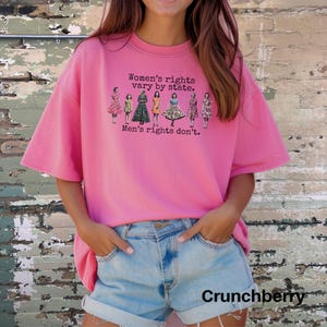 Puede incluir: Una camiseta rosa con el texto "Women's rights vary by state. Men's rights don't." e ilustraciones de mujeres con vestidos. La camiseta se combina con pantalones cortos vaqueros.