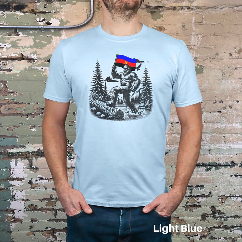 Peut inclure: T-shirt bleu clair avec une illustration en noir et blanc de Bigfoot tenant un drapeau rouge, bleu et noir. Le motif comprend des arbres, une b&ucirc;che et de l'eau. Le texte "Light Blue" est en bas.