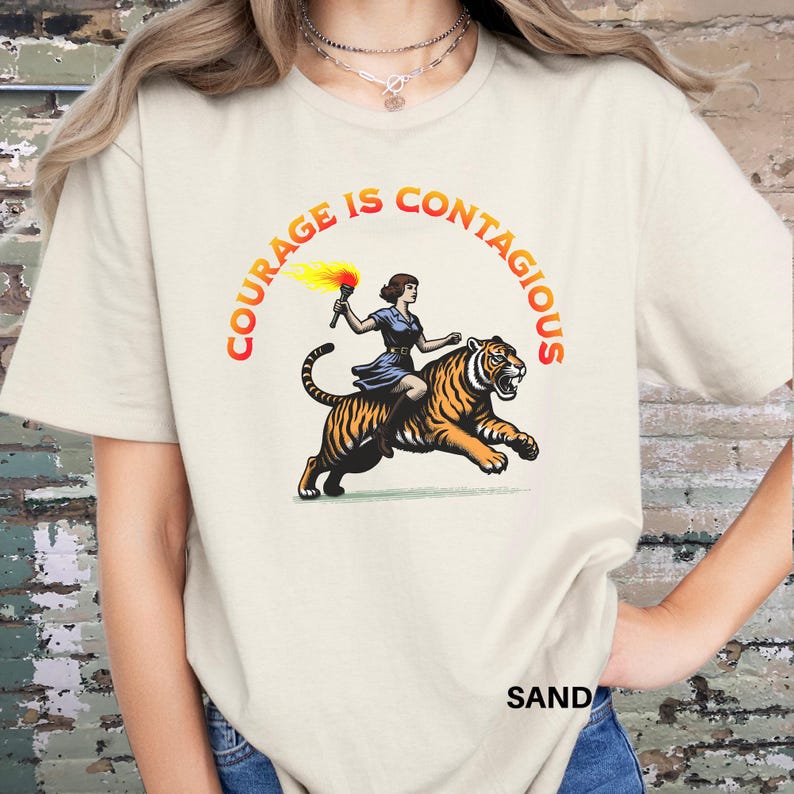 K&ouml;nnte beinhalten: Ein wei&szlig;es T-Shirt mit dem Text "Courage is Contagious" in orangefarbenen und gelben Buchstaben. In der Mitte des Shirts ist eine Frau abgebildet, die auf einem Tiger mit einer Fackel reitet.