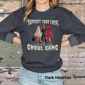 K&ouml;nnte beinhalten: Ein dunkelgrau meliertes Sweatshirt mit Rundhalsausschnitt und einem Grafikdruck von drei Figuren in Halloween-Kost&uuml;men sowie dem Text "SUPPORT YOUR LOCAL GHOUL GANG" in heller Schrift. Die Figuren zeigen ein Gespenst, ein Skelett und eine K&uuml;rbiskopf-Figur.