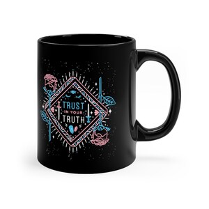 May include: Black ceramic mug with a white design featuring a diamond shape with a blue and pink outline. The diamond shape contains the text "TRUST IN YOUR TRUTH" with a sword, eye, and crystal graphic. The background is black with white stars.