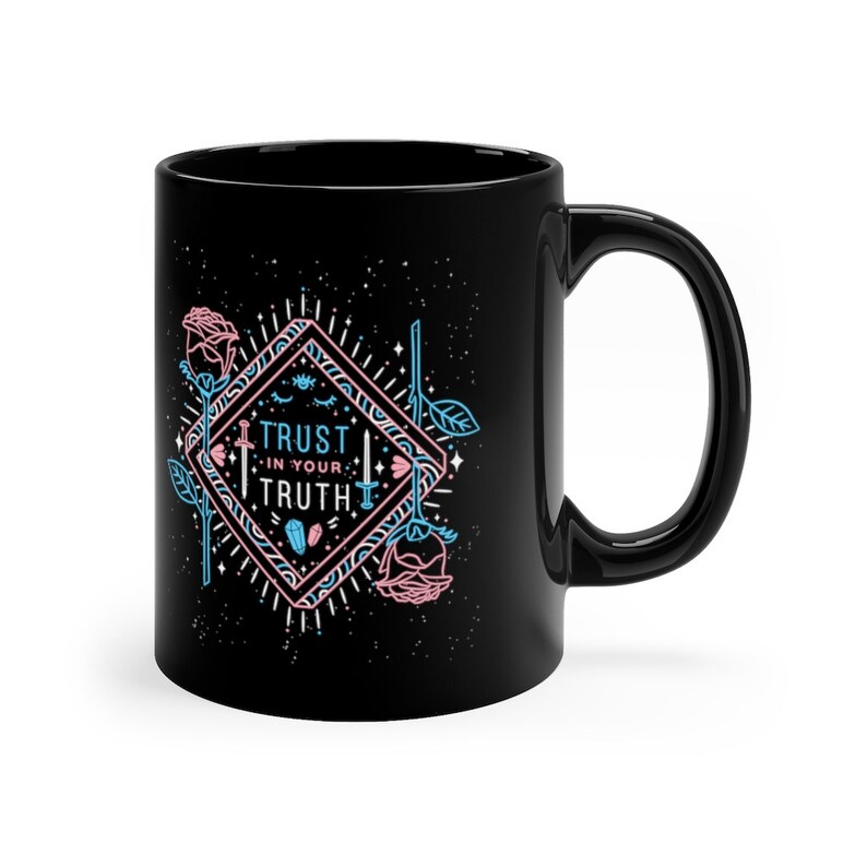 May include: Black ceramic mug with a white design featuring a diamond shape with a blue and pink outline. The diamond shape contains the text "TRUST IN YOUR TRUTH" with a sword, eye, and crystal graphic. The background is black with white stars.
