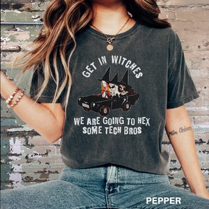 Może przedstawiać: Szary t-shirt z vintage'owym wyglądem z grafiką przedstawiającą trzy czarownice jadące samochodem z tekstem "Get in Witches We are going to hex some tech bros".