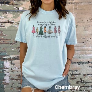 Puede incluir: Camiseta azul claro con el texto "Women's rights vary by state. Men's rights don't." El gr&aacute;fico presenta ilustraciones de mujeres con vestidos. La palabra "Chambray" est&aacute; impresa en la parte inferior de la camiseta.