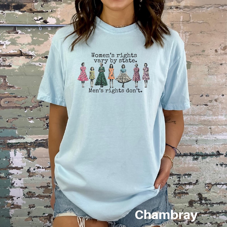 Puede incluir: Camiseta azul claro con el texto "Women's rights vary by state. Men's rights don't." El gr&aacute;fico presenta ilustraciones de mujeres con vestidos. La palabra "Chambray" est&aacute; impresa en la parte inferior de la camiseta.