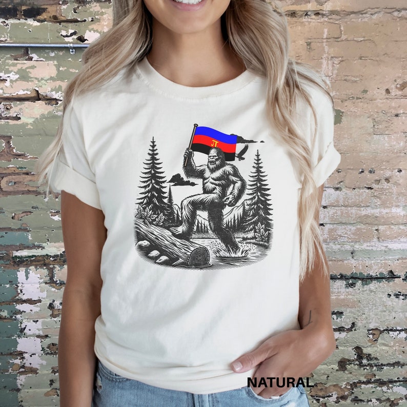 Peut inclure: T-shirt cr&egrave;me avec une illustration en noir et blanc de Bigfoot tenant un drapeau avec des rayures rouges, bleues et noires. Le motif comprend des arbres, une b&ucirc;che et le mot "NATURAL" en bas.