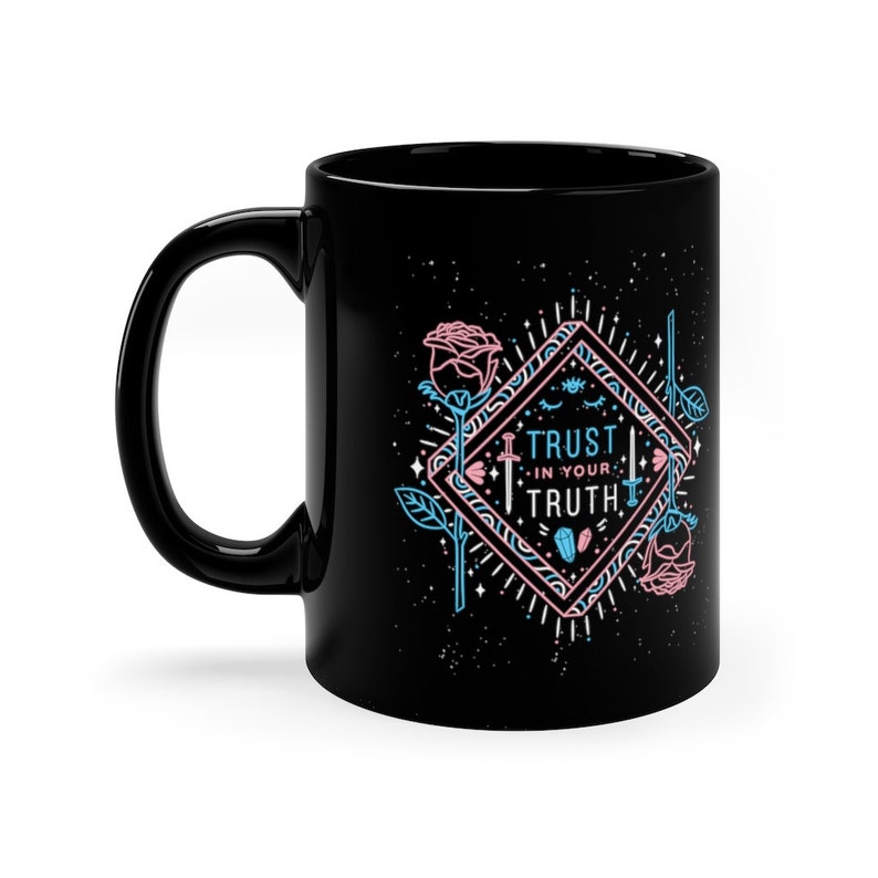 May include: Black ceramic mug with a white design featuring a diamond shape with the text "Trust in your Truth" in pink and blue lettering. The design also includes two pink roses, two swords, and stars.