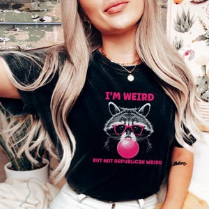 Peut inclure: T-shirt noir avec un raton laveur rose portant des lunettes de soleil roses et soufflant une bulle rose. Le t-shirt porte l'inscription "I'm Weird But Not Republican Weird".
